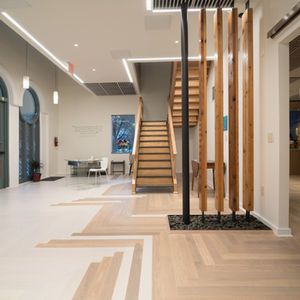 Spacious entry lobby and reception area at Kadampa Meditation Center in Historic Dilworth, Charlotte, NC after adaptive reuse renovation by Held Architecture. Features dramatic herringbone wood flooring, open staircase, custom wooden slat screen with pebble base, arched windows, and views into the illuminated meditation hall with golden Buddha statues.