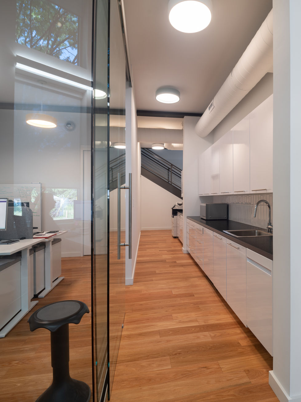 Modern kitchenette at VS America Office & Showroom in Charlotte, NC, featuring sleek white high-gloss cabinetry, black countertops with stainless steel sink, exposed white ductwork, circular ceiling lights, glass partition wall, black stool, and light wood flooring – part of the timeless modern renovation by Held Architecture.