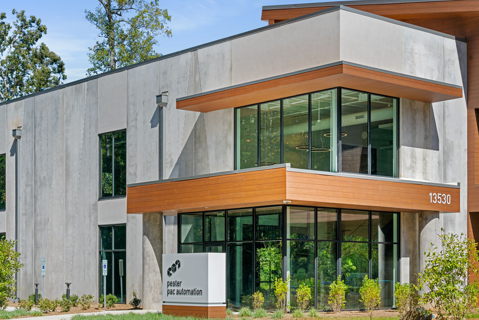 Close-up entrance detail of Pester Pac Automation USA Headquarters in Huntersville, North Carolina, designed by Held Architecture. Modern biophilic facade featuring exposed concrete panels, warm wood cladding, large glass windows, company signage, and elevated architectural elements.