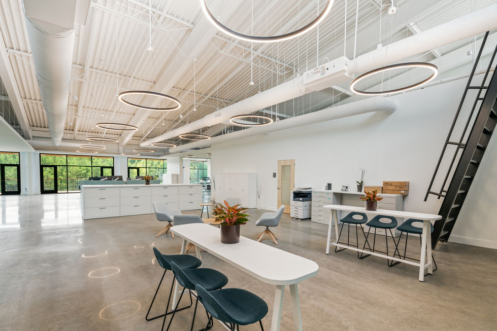 Bright collaborative breakout area at Pester PAC Automation USA headquarters in Huntersville, NC featuring high-top white tables with dark green chairs, exposed white ceiling structure and ductwork, circular pendant lights, polished concrete floors, and abundant natural daylight from large windows – biophilic design by Kristina Held.