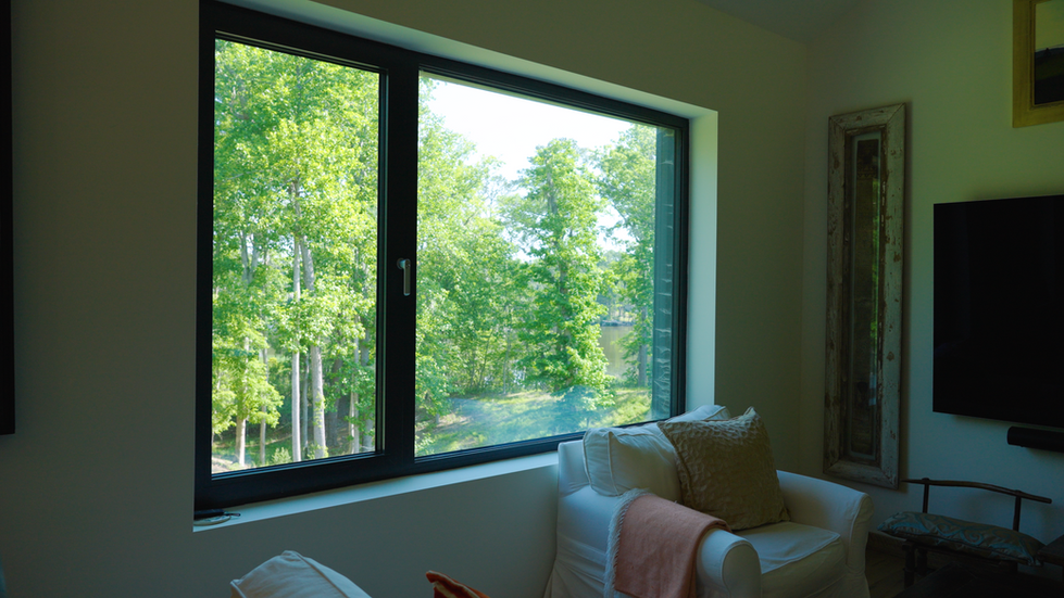 Interior living area of Nevills Creek Passive House showing a large picture window with stunning views of lush green forest and waterfront, cozy white armchair, and abundant natural light. Off-grid Passive House designed by Held Architecture in New Bern, North Carolina.