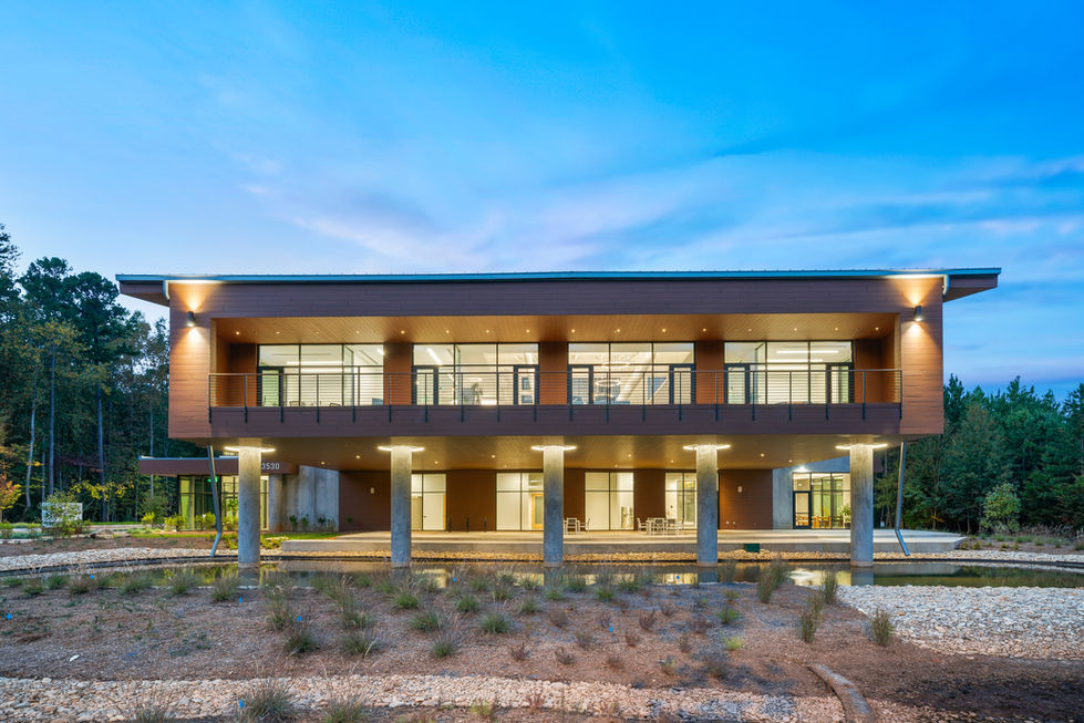 Front elevation dusk view of Pester Pac Automation USA Headquarters in Huntersville, North Carolina, designed by Held Architecture. Striking biophilic building with elevated wood-clad volume on concrete pillars over water feature, glowing interiors, exterior lighting, and deep connection to the forested landscape.
