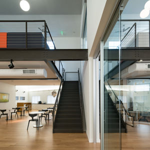 Modern double-height interior at VS America Office & Showroom in Charlotte, NC, featuring a prominent black central staircase, mezzanine with metal railings, exposed white ductwork, open collaboration areas with tables and colorful seating, all part of the timeless renovation by Held Architecture.