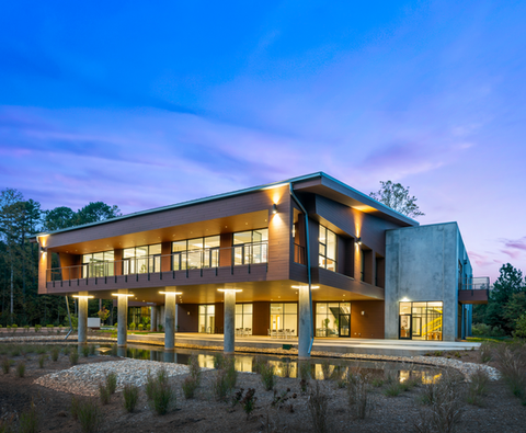 Wide dusk twilight view of Pester Pac Automation USA Headquarters in Huntersville, North Carolina, designed by Held Architecture. Dramatically illuminated biophilic building with elevated wood structure on concrete pillars over water feature, glowing interiors, and stunning colored sky reflecting in the pond.