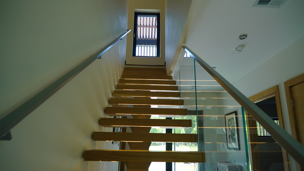 Dramatic low-angle view of the modern interior staircase at Nevills Creek Passive House, featuring warm wood treads with integrated LED lighting, sleek glass railing, metal handrail, and natural light from the upper window overlooking the forest. Off-grid sustainable home designed by Held Architecture in New Bern, North Carolina.