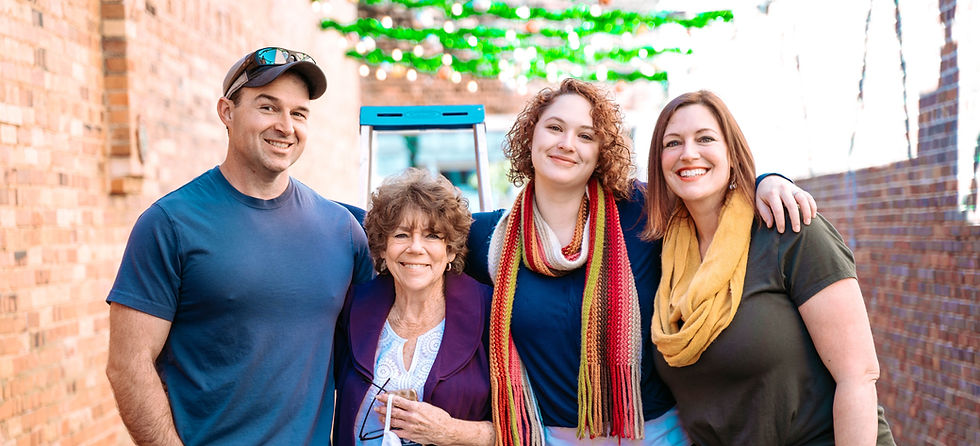 Four people smiling in front of a brick wall. Bright scarves and shirts add color. Green lights and ladder in the background. Happy mood.