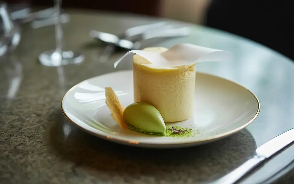 Soufflé glacé individuel pastel (vanille/pistache) avec collerette en papier, voile de sucre glace, servi en dessert léger sur assiette blanche minimaliste avec quenelle de sorbet, micro-herbes et éclats de meringue, table en marbre clair, ambiance restaurant étoilé en bokeh — Les Saveurs de René. Negative prompt : texte, typographie, lettres, mots, logo, watermark, étiquette, menu imprimé, marque, packaging, artefacts, mains, visage, personnage, fond encombré, couleurs criardes, style cartoon, illustration plate.