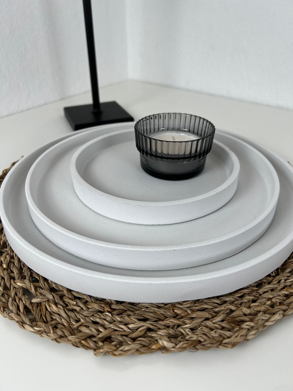 Weiße Betontabletts in drei Größen mit Teelicht dekoriert und Lampe im Hintergrund auf einem geflochtenem Untersetzer