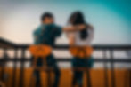 A young couple sits on high stools at a rooftop bar or terrace, facing the horizon. The man, wearing a patterned shirt, has his arm around the woman, who is dressed in a white top and jeans. The sky displays a gradient of warm and cool tones, creating a relaxed and intimate atmosphere