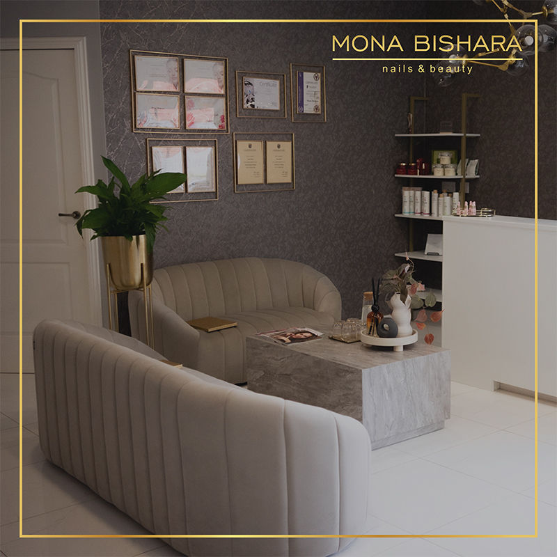 A cozy salon waiting area with beige sofas, a gray coffee table, plants, and wall certificates. Shelves hold beauty products. Warm atmosphere.