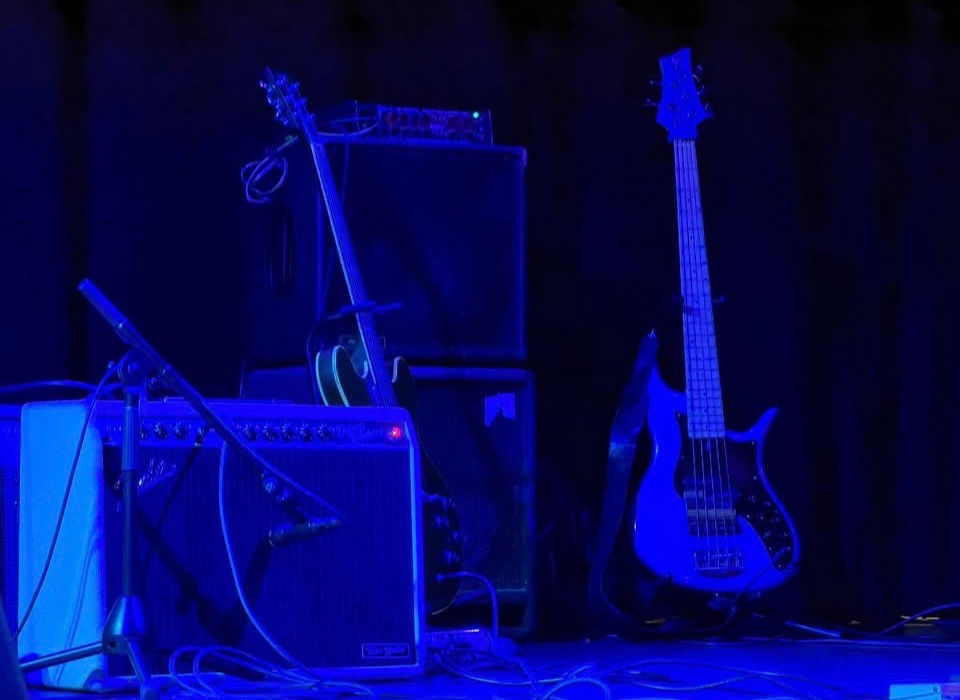 A close-up shot of an electric bass guitar standing next to stage amplifiers and a microphone stand under deep blue stage lighting.