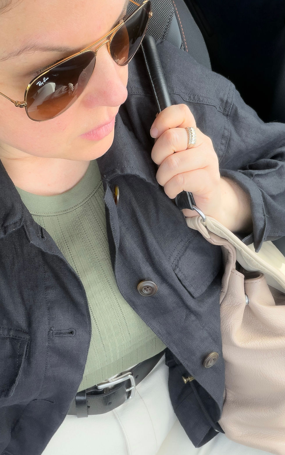 Personal styling feature by Michelle Marie Sawtell: A high-angle selfie detailing a curated outfit that demonstrates intentional metal mixing. Michelle wears gold aviator sunglasses and a gold ring alongside a black leather belt featuring a prominent silver buckle. Her look is completed with a green pointelle knit top, a black button-down linen jacket, and a neutral beige American Leather Co. handbag.