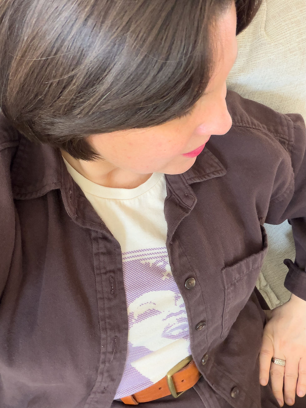 A high-angle style detail photo by Michelle Marie Sawtell highlighting a monochromatic brown wardrobe look. Michelle models an open dark chocolate brown denim button-up shirt worn over a cream graphic t-shirt featuring a muted purple illustration. The outfit is cinched with a warm maple-toned leather belt with a brass buckle at the waist of matching brown denim jeans.