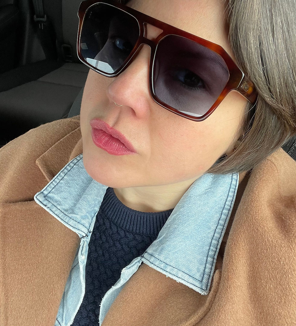 A close up profile portrait of Michelle Marie Sawtell inside a vehicle emphasizing the fine details of her eyewear and layered garments. The camera captures her face from a side angle, showcasing the gold brand emblem on the tortoiseshell temple arm of her Quay navigator sunglasses. The light wash denim shirt collar features clean stitching along its border, layered over a dark cable knit sweater and enveloped by a plush camel wool coat. Her hands are completely out of the frame.
