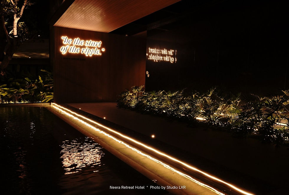 Neon quote "be the start of the ripple" glows on a dark wood wall, with plants below. A pool reflects the light, creating a serene mood.