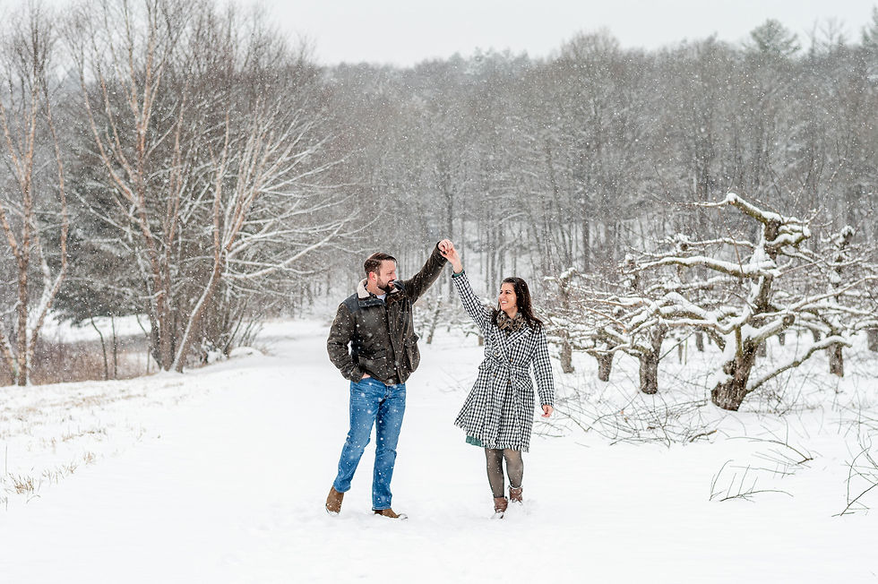 Couple dancing in a snowy forest during engagement portraits, with bare trees and snow falling. They appear joyful, wearing winter coats, in a serene, wintry landscape.