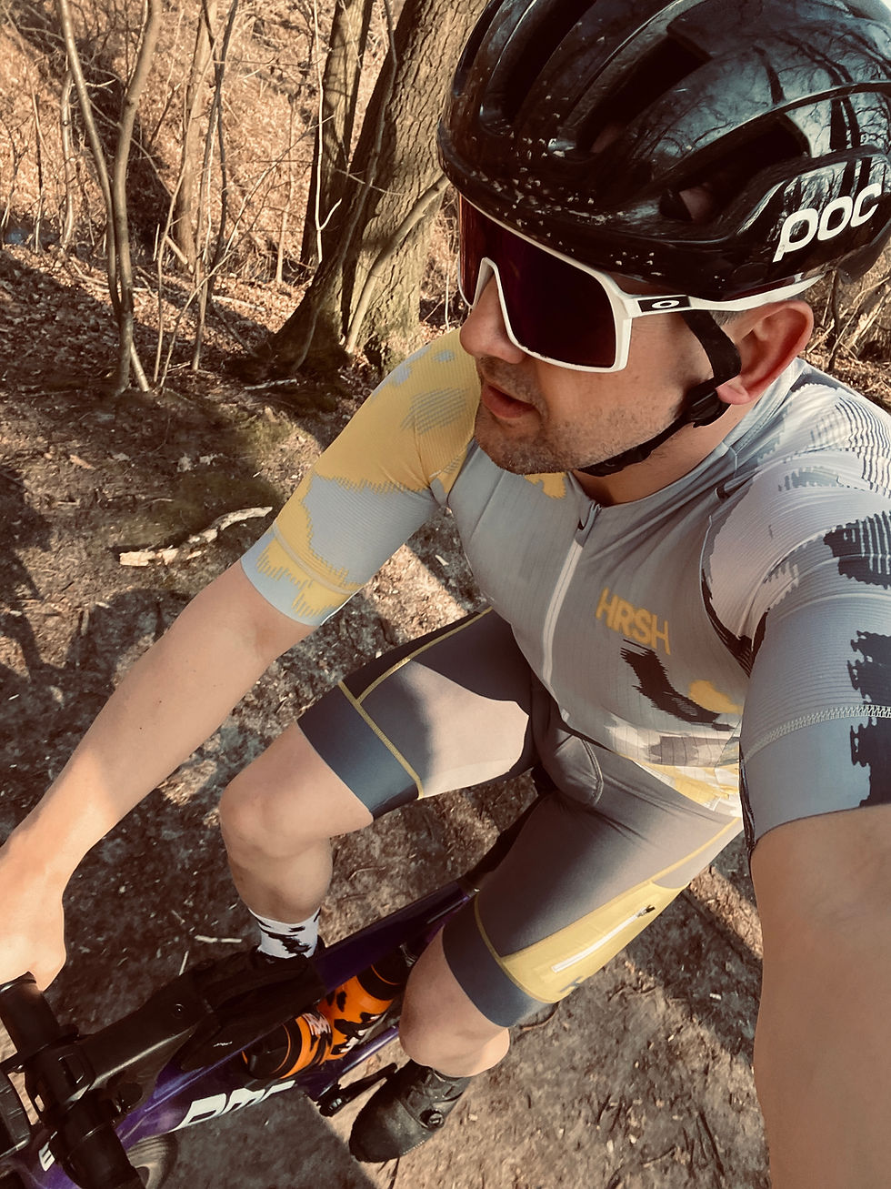 Cyclist in gray and yellow kit rides a bike on a forest trail. Wearing a black helmet and sunglasses. Sunlit trees surround the path.