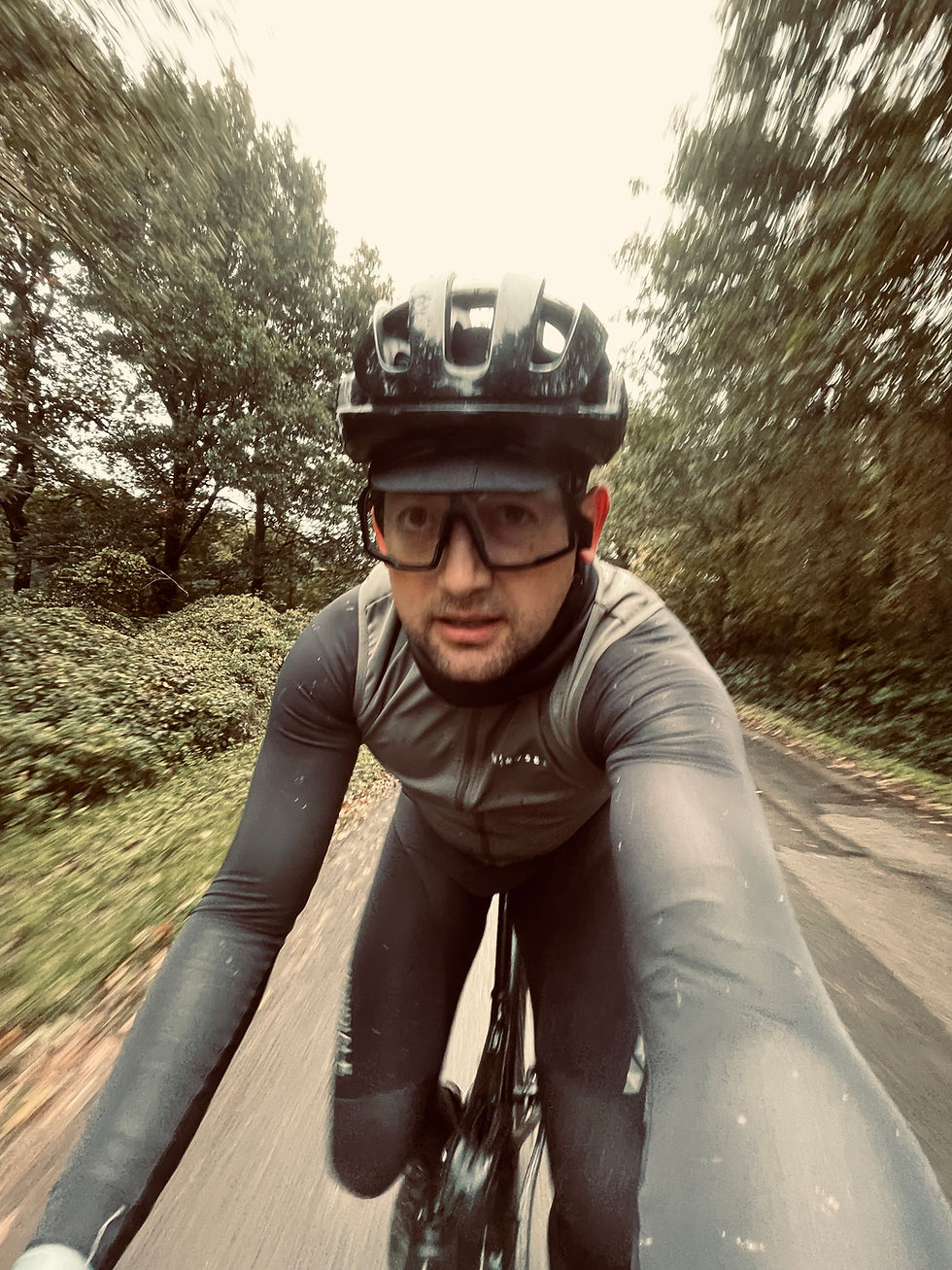 Cyclist in helmet and glasses riding on a forest road, wearing a gray outfit. Trees blurred in motion, overcast sky. Determined expression.