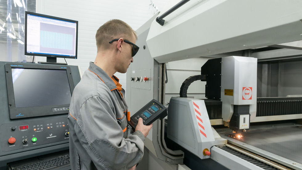 An industrial operator wearing safety glasses and a grey work jacket stands before a high-precision laser cutting machine in a clean, modern factory. He holds a handheld digital control unit to monitor active processing conditions while a spark indicates the machine is in operation. To his left, a control station with multiple monitors displays process data, illustrating the integration of technical expertise and data-driven condition management.