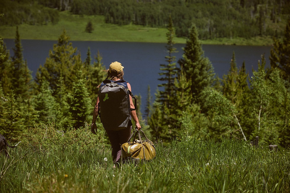 Hiker carrying Eagle Creek Migrate duffel bag and backpack through alpine meadow toward a lake.