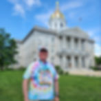 Pete in a tie-dyed shirt, standing in front of the NH Statehouse