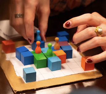 Main BLCS project image. The image shows a small square game board made of cardboard and paper, with colorful blocks and pawns placed on top of it. A hand in the background moves a blue pawn atop a blue block on the board, while another hand reaches from the right side of the image towards the center blocks.