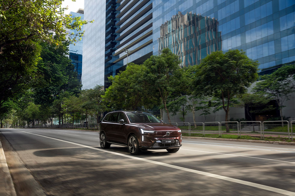 SUV marrom Volvo EX90 em avenida arborizada, com prédios de vidro refletindo a cidade.