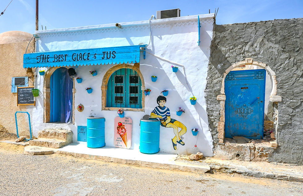 Colorful café façade in Djerba, Tunisia, with whitewashed walls, bright blue doors and barrels, flower pots, and a mural of a boy sitting beside a painted ice cream sign under a blue awning reading “The Best Glace & Jus.”