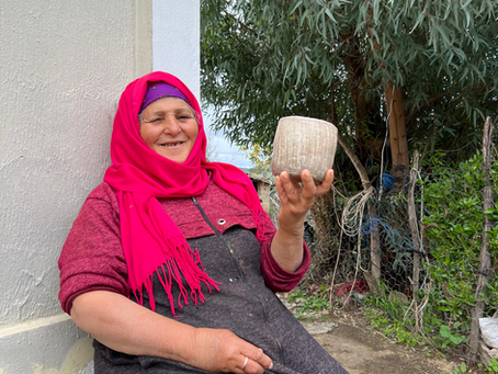 Chmissa, a Tunisian artisan potter from Sejnane, seated in her courtyard holding a handmade clay vessel with a smile.