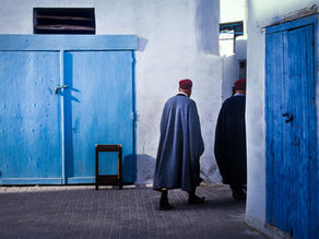Two men walking through a quiet street lined with blue doors and whitewashed buildings in Tunisia