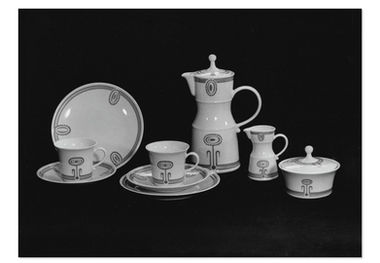 Schwarz-Weiß-Fotografie eines mehrteiligen Porzellan-Kaffeeservices, dem „Déjeuner“ für zwei Personen von Ellinor Symmangk aus dem Jahr 1973 für das Wissenschaftlich-Technische Zentrum der Feinkeramik (WTK) Meissen. Das Service steht auf einer dunklen Oberfläche vor schwarzem Hintergrund. Es besteht aus einer schlanken Kaffeekanne mit Deckel und Henkel, zwei Tassen auf passenden Untertassen, einem kleinen Milchkännchen, einer Zuckerdose mit flachem Deckel und einem größeren Teller im Hintergrund. Alle Teile sind aus weissem Porzellan gefertigt und mit einem markanten, von Ellinor Symmangk entworfenen Dekor verziert.