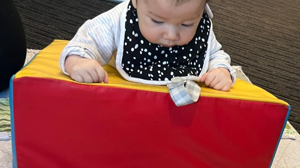Baby leaning on a red and yellow foam block, wearing a black bib with white dots. The baby is on a patterned mat in a carpeted room.