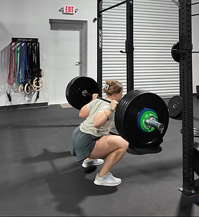 Woman performing a barbell squat in a gym. Surroundings include gym equipment and a closed door with an exit sign. Focused mood.