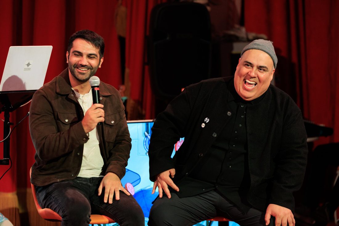 Adam with a microphone, laughing on stage beside composer Ego Plum