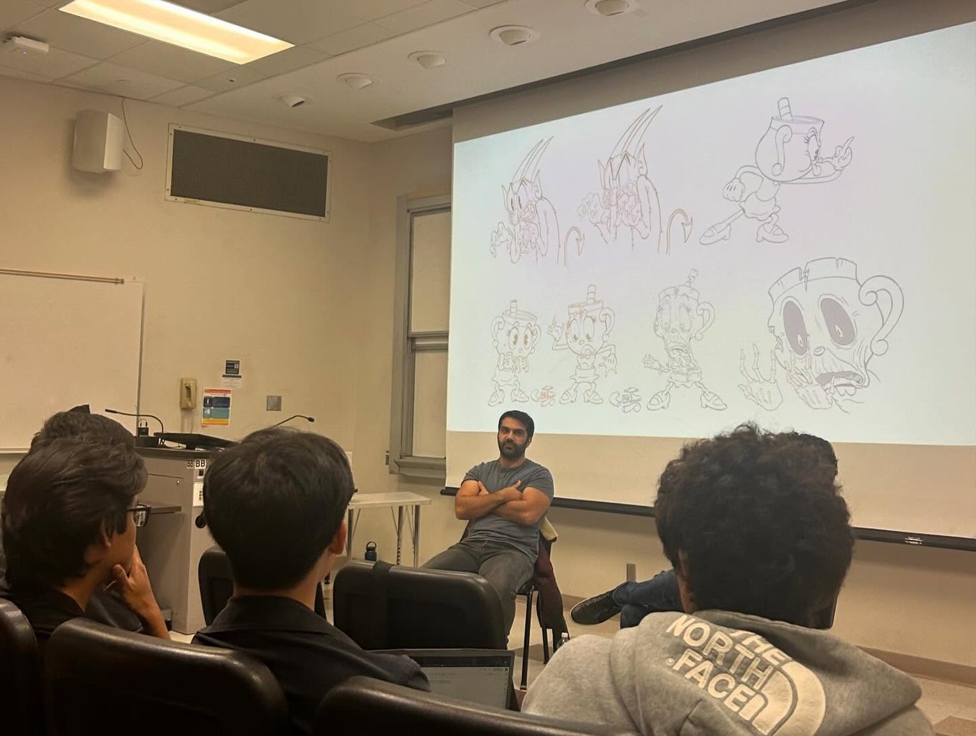 Adam seated before a lecture hall screen of his Cuphead concept drawings, students listening