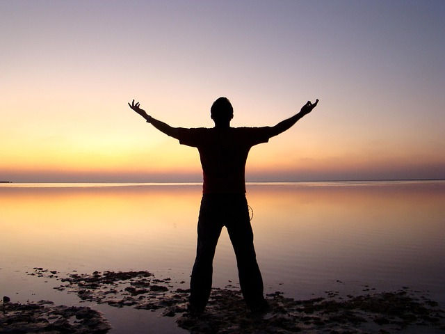 Silhouetted person with arms raised stands by calm water during a vibrant orange and purple sunset, conveying a sense of freedom.
