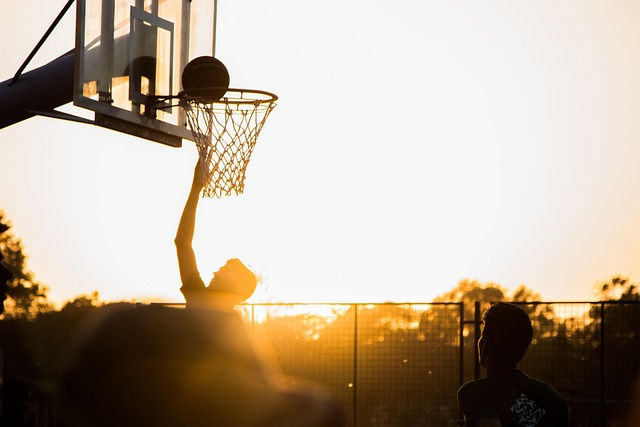Silhouetted basketball player jumps to score at sunset on an outdoor court, with trees in the background. Warm and energetic mood.