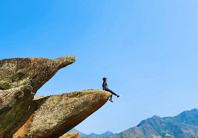 person sitting on the edge of a cliff overlooking some mountains with clear blue sky in background