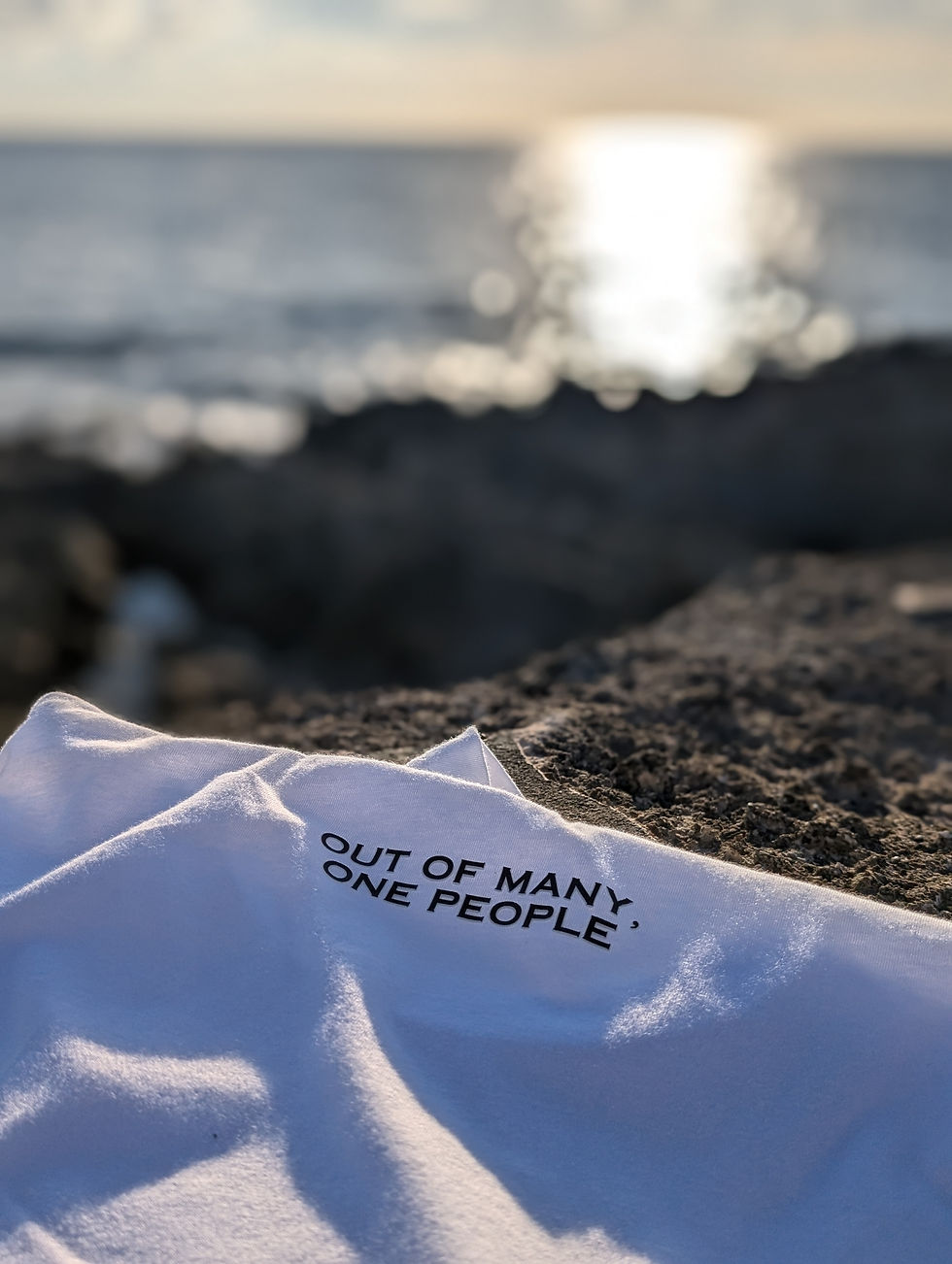 Thumbnail: White T-shirt with “Out of Many, One People” text lying on rocky shore at sunset, with golden light reflecting on the ocean.