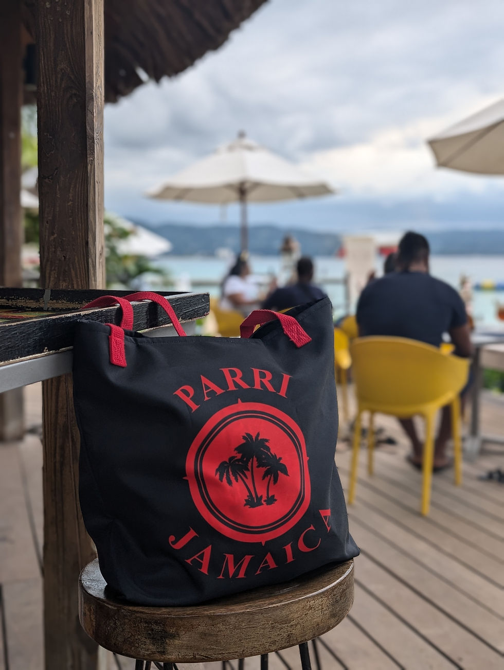 Thumbnail: Black beach tote with bold red “Parri Jamaica” logo and straps, on a beach bar stool with ocean background.