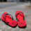 Thumbnail: Red Parri Jamaica flip flops with black straps on a large rock with the streets of Kingston Jamaica in the background.