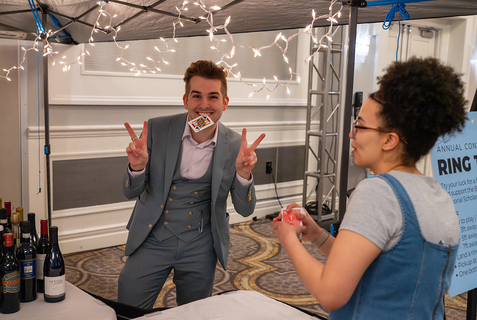 Brenden Trojan in gray suit flashes peace signs with a playing card in his mouth at a fairy-lit ring toss booth, while a woman holds cards.
