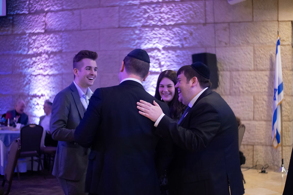 Four people in formal wear converse and smile in a warmly lit room with stone walls and an Israeli flag. The mood is joyful.