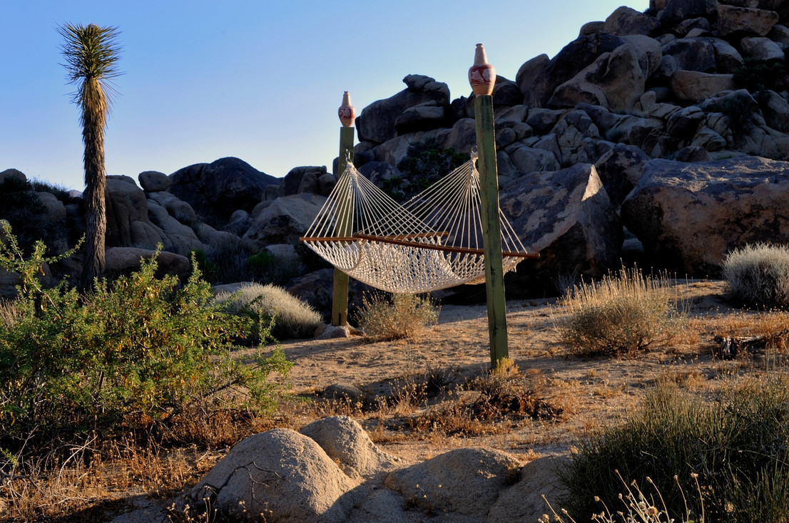 A white rope hammock strung between two wooden posts nestled among large desert boulders and shrubs.