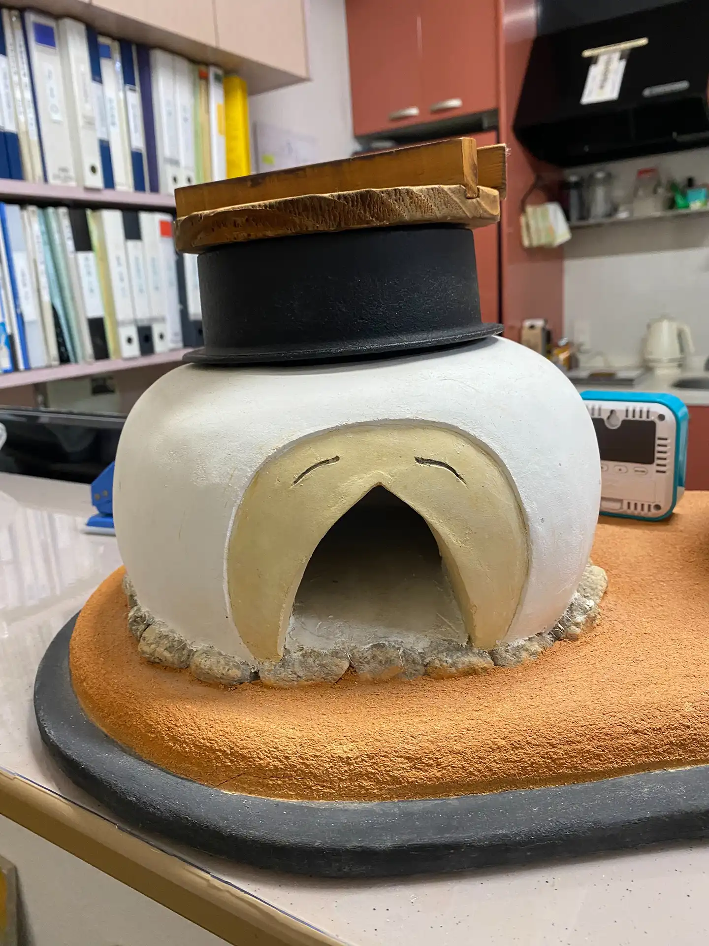 製作完成的白色圓潤土灶模型，開口處修飾為平滑圓弧，並帶有逗趣的表情刻紋，底部墊以橘色塗り版築底座。