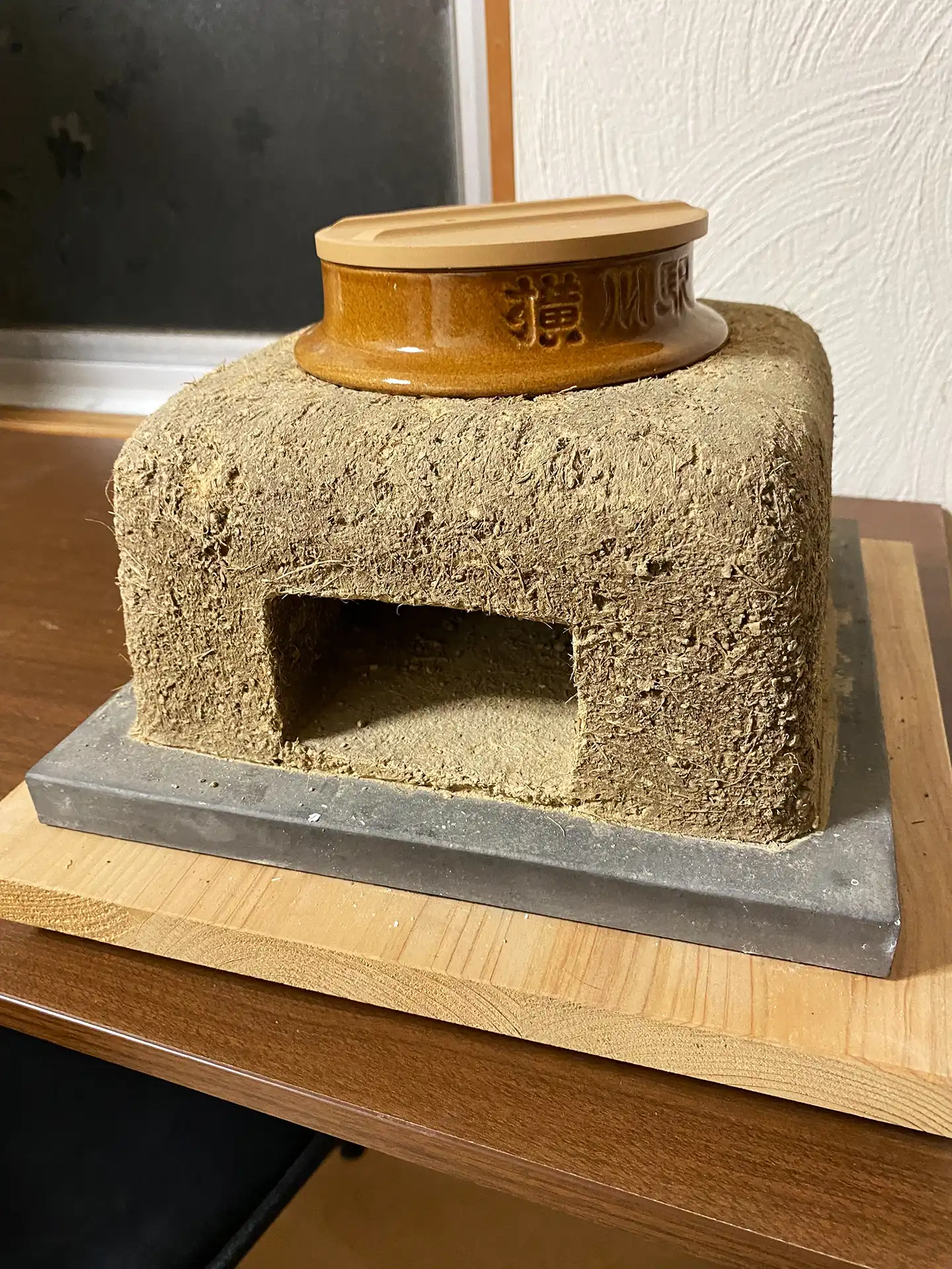 一個以粗質土壤與大量稻草纖維混合製成的烤爐（灶）雛形，上方放置咖啡色陶瓷鍋蓋，展示抹灰工藝初期的結構美感。