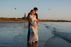 A pregnant woman with her partner hugging her from behind at sunrise on Longsands Beach in Tynemouth with St Georges church in the background_maternity photography by Emma Solomon Photography