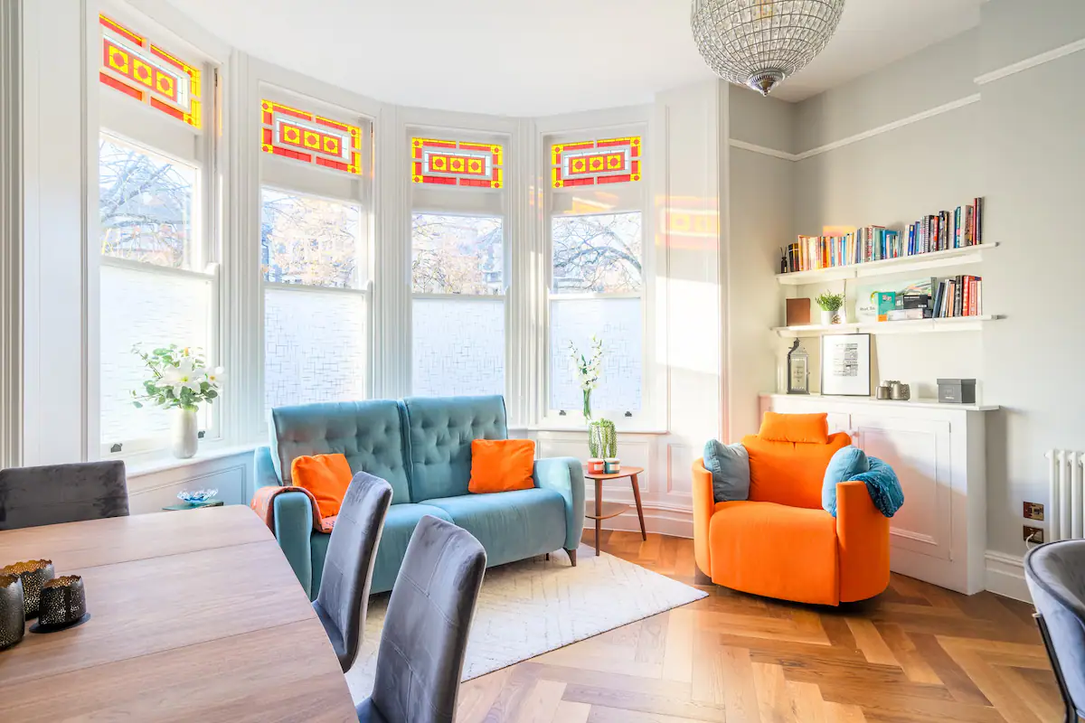 brightly lit living room with a blue and orange patterned couch. Natural light fills the room, highlighting the warm wood floors and colorful throw pillows