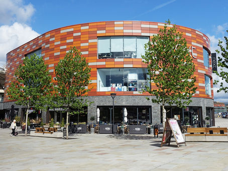 Modern shopping centre at the Friar's Walk in Newport with orange-tiled building, trees, and storefronts. People walking in sunny plaza. Signs for H&M and Next visible.