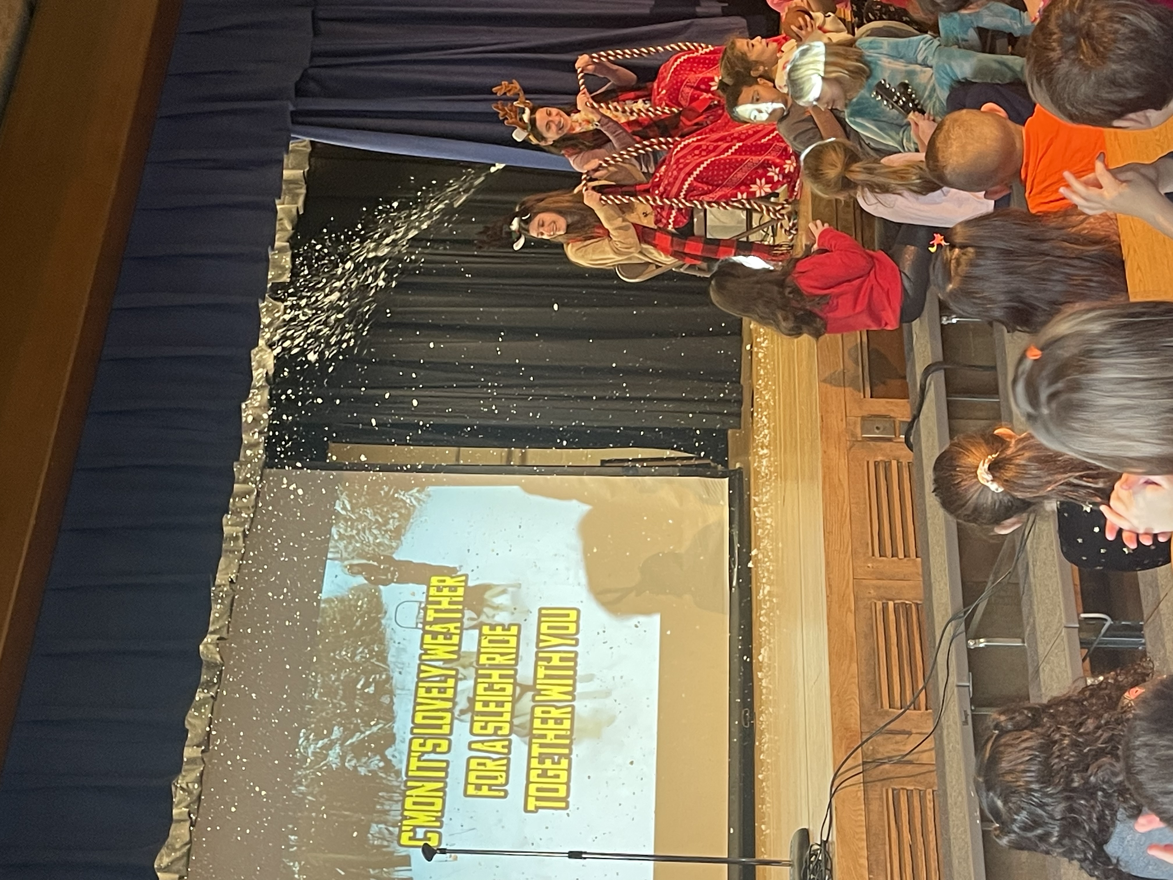 Elementary teachers dancing in a holiday-themed school assembly with indoor snowfall and live music